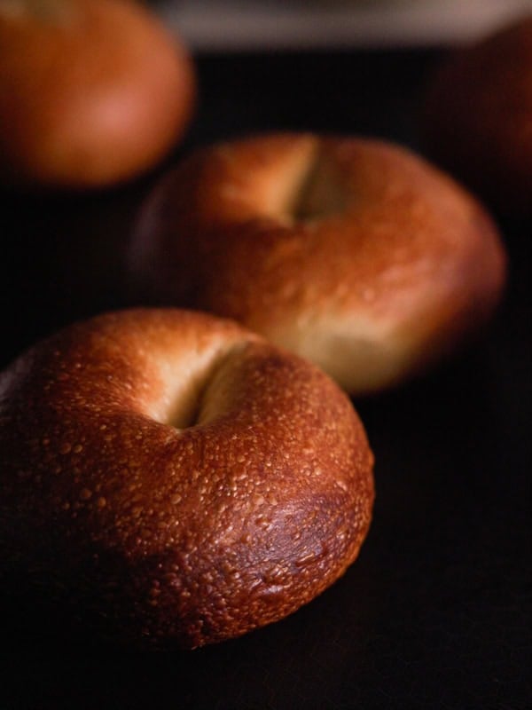 How to Make Delicious NY Style Bagels - Step by Step Guide. 6 Two golden brown NY style bagels with a slightly crisp texture sit on a dark surface, with blurred bagels in the background.