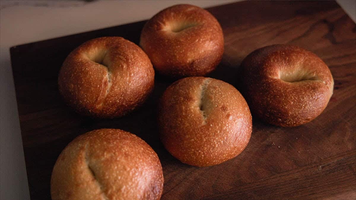 How to Make Delicious NY Style Bagels - Step by Step Guide. 2 Five golden-brown NY style bagels sit on a dark wooden cutting board, each with a slightly shiny crust and a small split or indentation in the center.