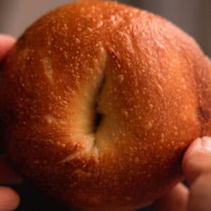 How to Make Delicious NY Style Bagels - Step by Step Guide. 100 Two hands hold a golden-brown bagel with a slightly shiny, textured crust and a visible split in the center, capturing the classic look of NY style bagels.