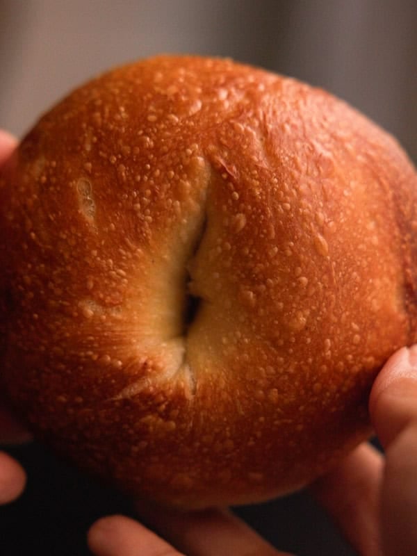 How to Make Delicious NY Style Bagels - Step by Step Guide. 79 A close-up of hands holding a golden brown, round bagel with a textured crust and a slight indentation in the center, showcasing the classic appeal of NY style bagels.