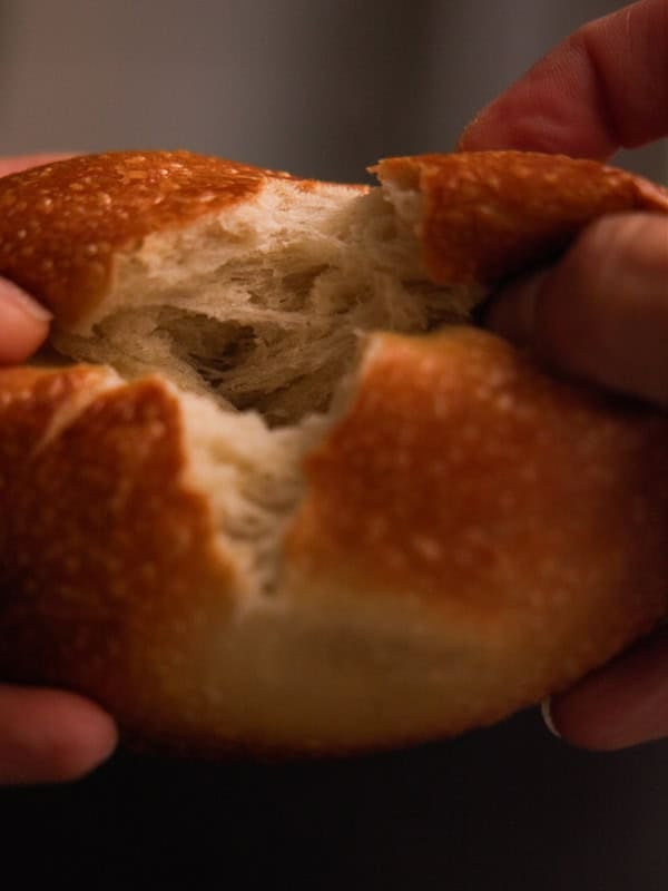 How to Make Delicious NY Style Bagels - Step by Step Guide. 86 A close-up of two hands tearing apart a round, golden-brown piece of bread, revealing its soft, airy interior—reminiscent of the irresistible texture found in NY style bagels.