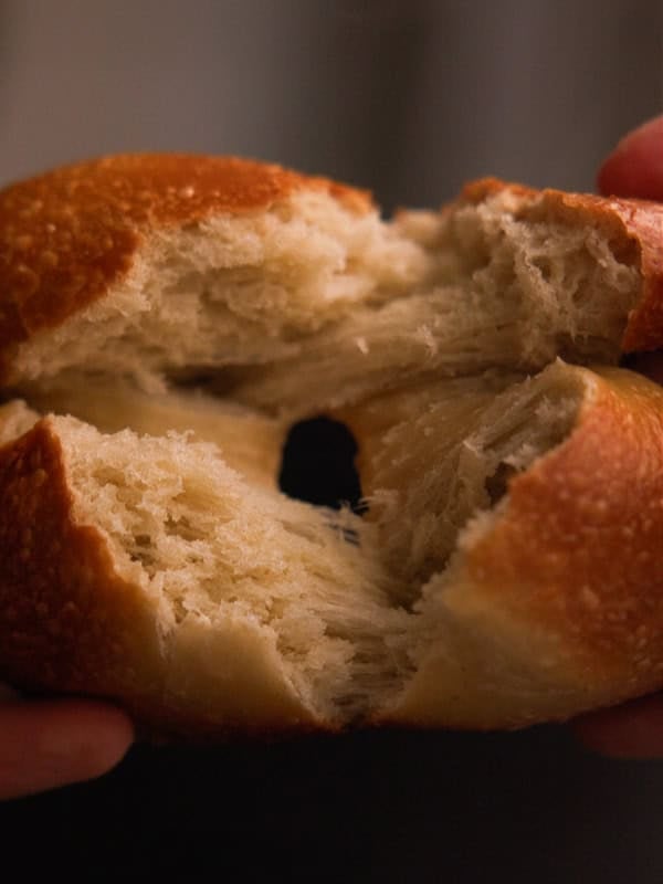 How My Bagels Triggered The Sexist In Me. 7 A close-up of hands pulling apart a NY style bagel, revealing its fluffy, soft interior and golden brown crust.