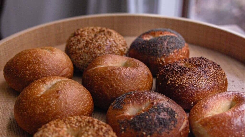 A wooden tray filled with nine NY style bagels, some topped with sesame seeds, some with poppy seeds, and others plain. A blurred window and curtain are in the background.