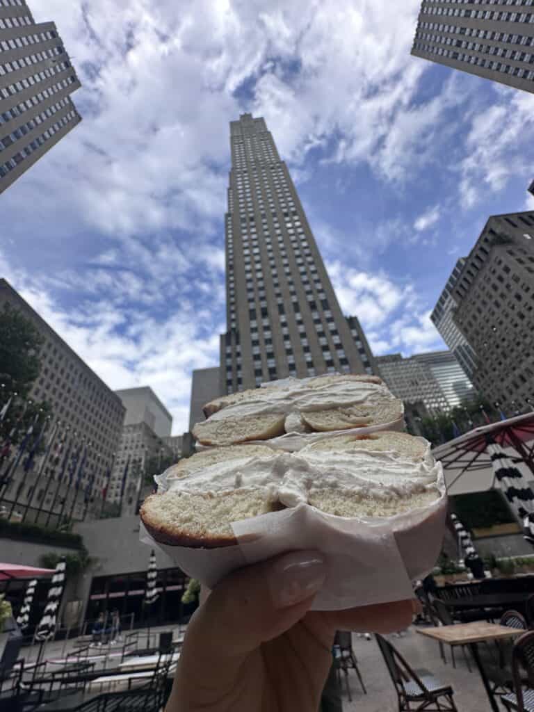 How to Make Delicious NY Style Bagels - Step by Step Guide. 5 A hand holds a classic NY style bagel with cream cheese in front of a tall skyscraper under a partly cloudy sky at Rockefeller Center, New York City.
