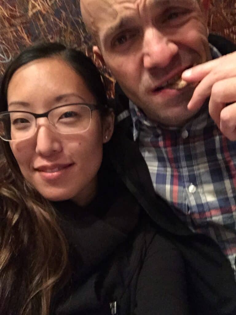 How to Make Delicious NY Style Bagels - Step by Step Guide. 4 Two people sit closely together. The woman on the left wears glasses and smiles. The man on the right, enjoying a classic NY style bagel, wears a plaid shirt and bites into his food with a playful expression. The background is blurred with warm tones.