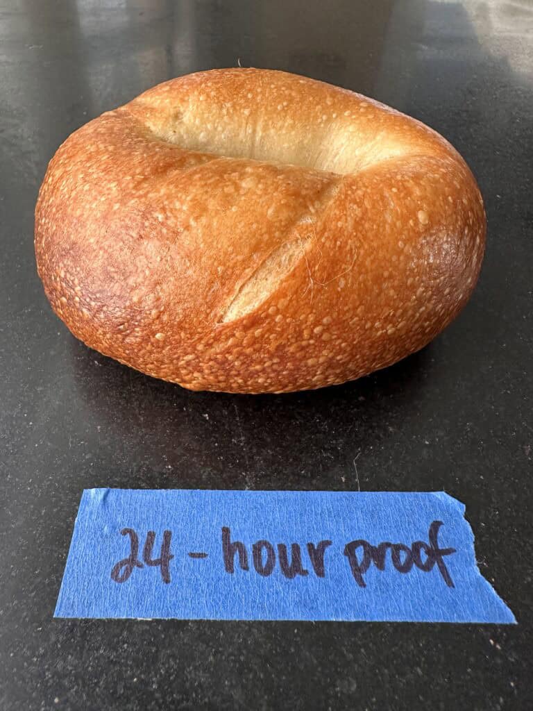 How to Make Delicious NY Style Bagels - Step by Step Guide. 81 A golden-brown NY style bagel sits on a dark surface with a piece of blue tape in front labeled “24-hour proof,” handwritten in black marker.