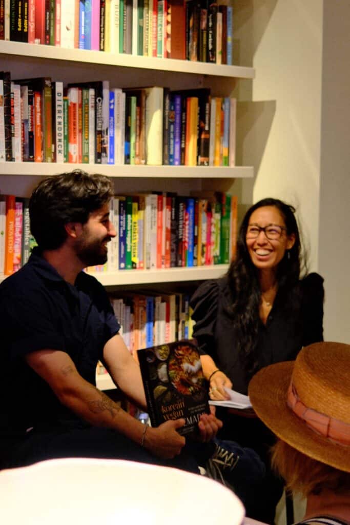How My Bagels Triggered The Sexist In Me. 11 Two people sit and smile in front of a bookshelf, sharing a personal story. One holds a book titled Korean Vegan, and the friendly setting is enhanced by the casual presence of bagels on the table. A third person in a hat is in the foreground, partially visible, adding to the warm and relaxed atmosphere.
